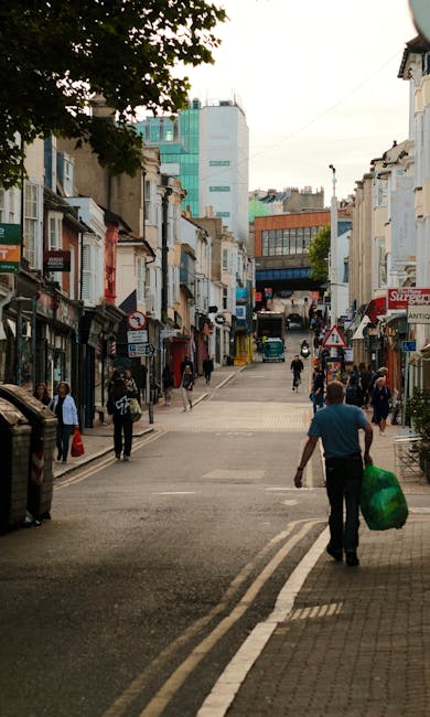 A busy urban street scene in Wimbledon Village with a mix of pedestrians walking along the pavement and crossing the road, some carrying shopping bags or backpacks. The foreground features a man wearing a blue shirt and dark trousers, holding a large green plastic bag filled with rubbish or discarded items. To the left, a woman carries a shopping bag, while behind her others continue their walk or wait at the crossing. The street is lined with small retail shops and cafes with signage, awnings, and shopfronts in various colors, including white, black, and red. On the right, a sidewalk displays potted plants and outdoor seating areas. The background reveals a slight incline with buildings of different architectural styles and heights, including older masonry structures and modern glass-fronted buildings, with some construction or renovation activity visible further along the street. The sky appears overcast, casting muted natural light over the scene, which also subtly indicates the importance of local waste collection and disposal services like those provided by House Clearance Wimbledon for maintaining cleanliness in such busy public areas.