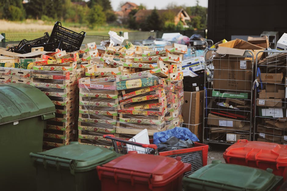 An outdoor scene showing a collection of piled cardboard boxes and plastic crates filled with assorted packaging materials, including fruit box cartons with colorful printed labels and black plastic crate stacks. The boxes are stacked on a paved surface, with some boxes slightly disarranged and leaning against each other. In the foreground, multiple large plastic wheelie bins in green and red are visible, partially blocking the view of the boxes. To the right, there are metal wire shelves holding additional cardboard boxes and packaging supplies, some with labels indicating recycling or storage. The background features a blurred residential area with green trees and a few houses, under natural daylight. The scene suggests a private waste or rubbish collection setup, consistent with independent rubbish removal or on-site clearance services as offered by House Clearance Wimbledon, for efficient disposal or collection of rubbish materials.
