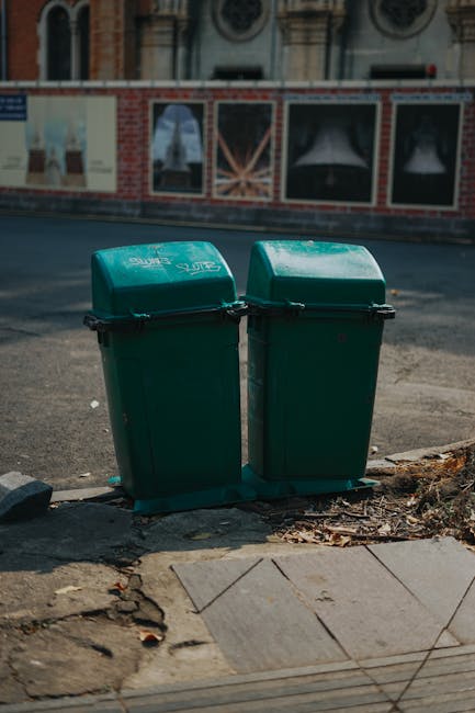 An outdoor scene showing a collection of piled cardboard boxes and plastic crates filled with assorted packaging materials, including fruit box cartons with colorful printed labels and black plastic crate stacks. The boxes are stacked on a paved surface, with some boxes slightly disarranged and leaning against each other. In the foreground, multiple large plastic wheelie bins in green and red are visible, partially blocking the view of the boxes. To the right, there are metal wire shelves holding additional cardboard boxes and packaging supplies, some with labels indicating recycling or storage. The background features a blurred residential area with green trees and a few houses, under natural daylight. The scene suggests a private waste or rubbish collection setup, consistent with independent rubbish removal or on-site clearance services as offered by House Clearance Wimbledon, for efficient disposal or collection of rubbish materials.